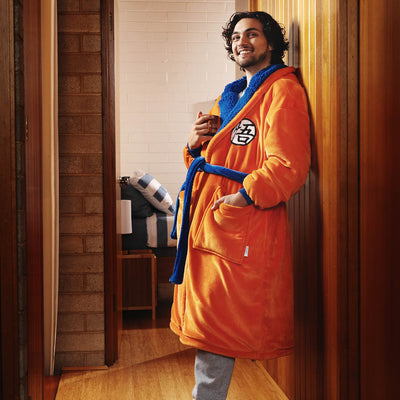 Person smiling and leaning against a wooden wall, wearing an orange and blue Dragon Ball Super-themed robe with a symbol on the chest pocket area.