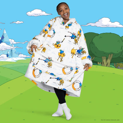 Smiling person wearing a white oversized wearable blanket covered in Adventure Time characters, standing on a green field with cartoon trees and mountains in the background.