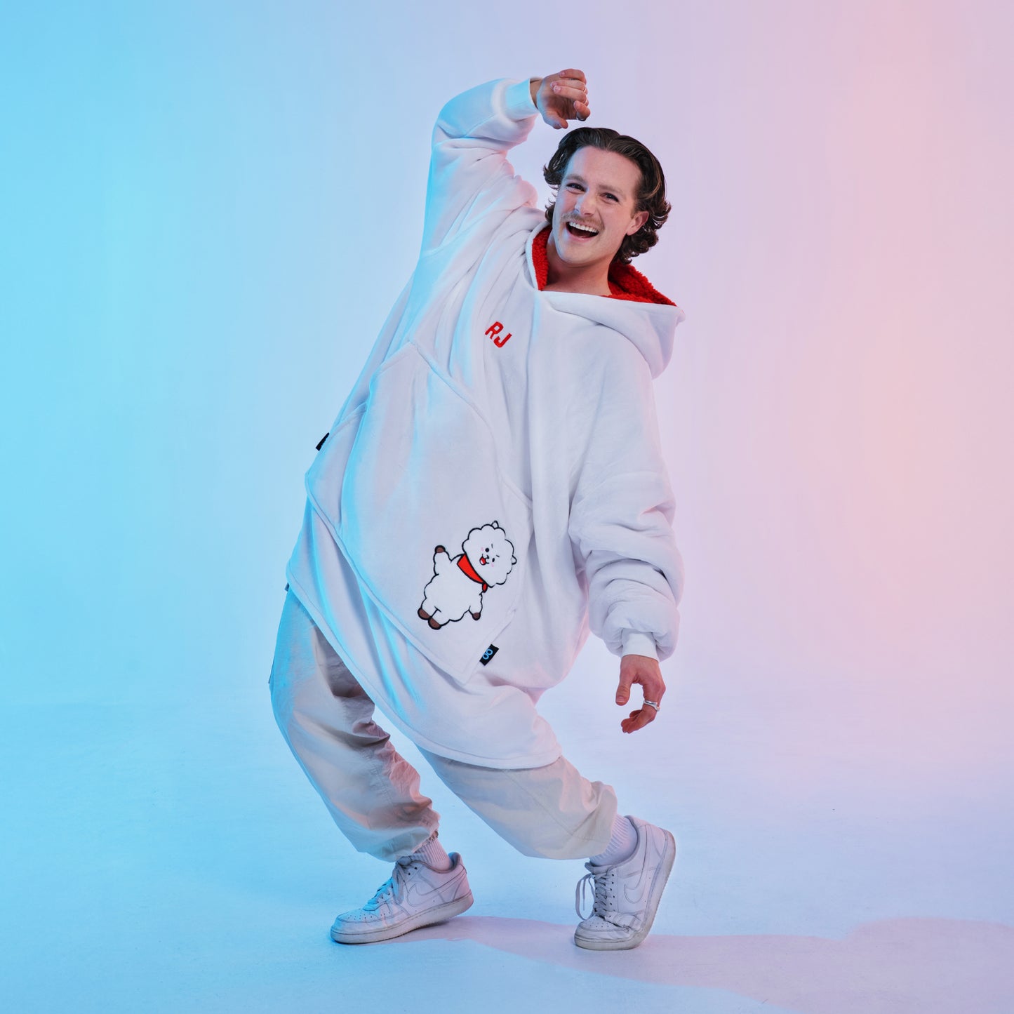Person in white oversized hoodie with cartoon dog and letters 'RJ', posing against blue and pink gradient background