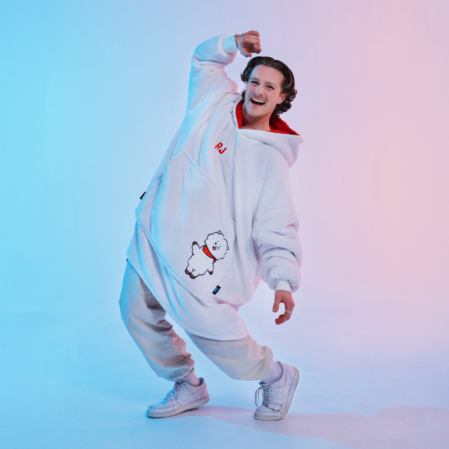 Person in white oversized hoodie with cartoon dog and letters 'RJ', posing against blue and pink gradient background