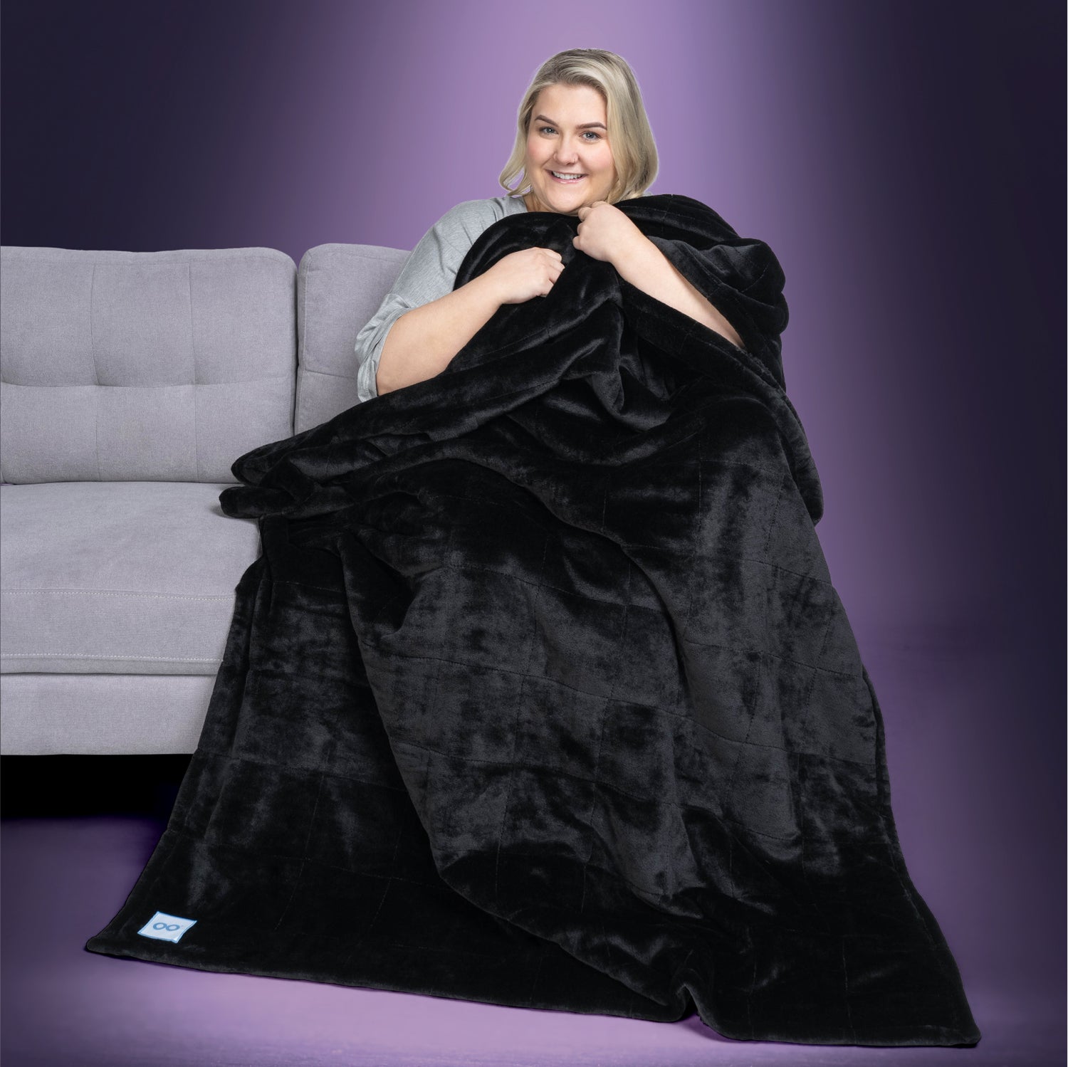 Woman sitting on a gray couch wrapped in a large black plush blanket with a small white tag visible at the bottom corner.
