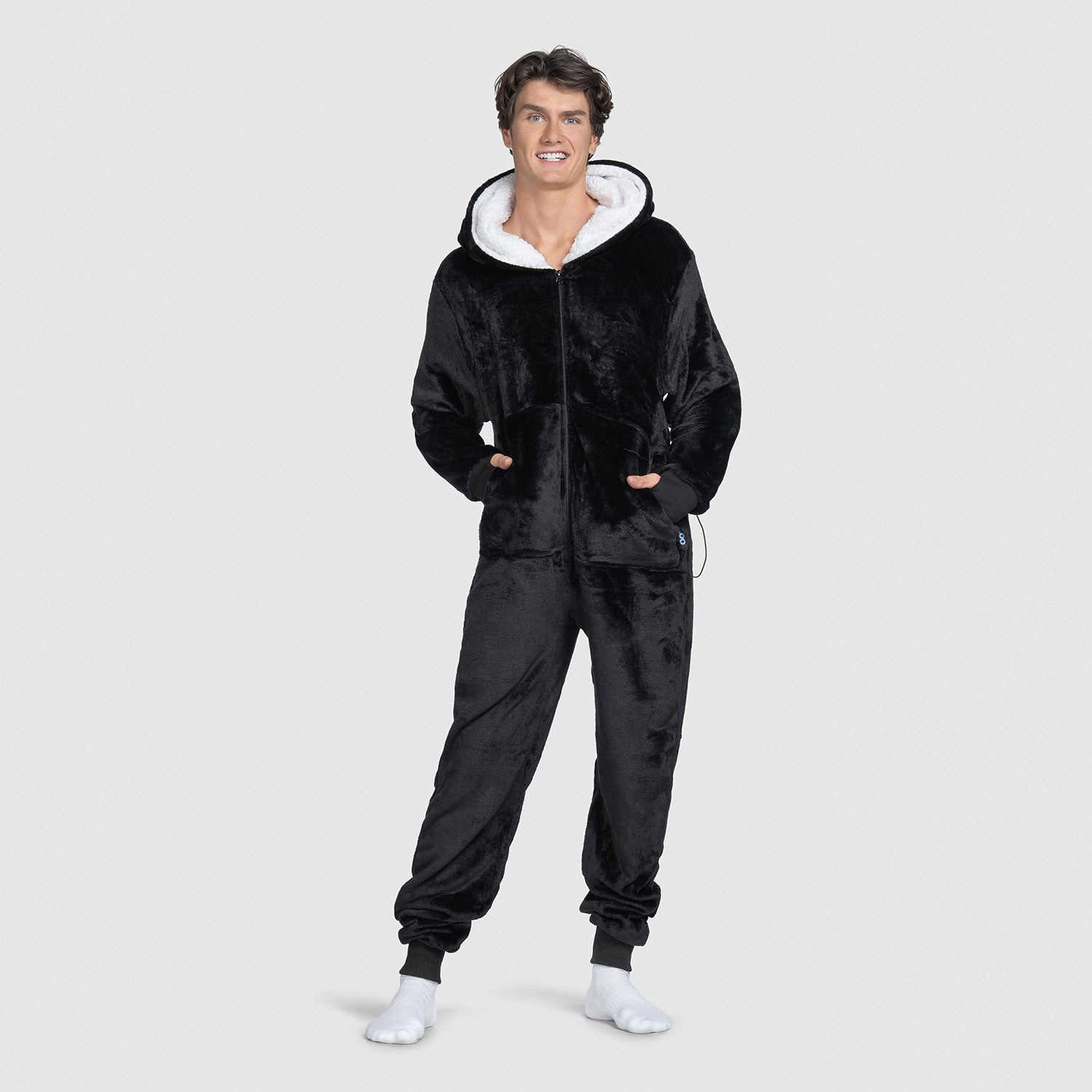 Man wearing a black plush hooded onesie with white lining, standing with hands in pockets and smiling.