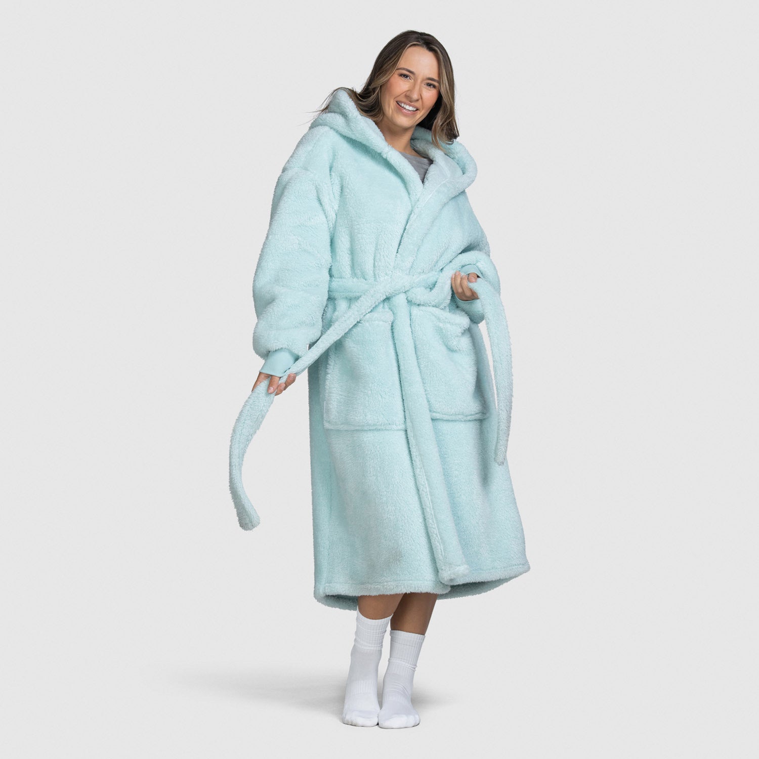 Woman wearing a light blue hooded bathrobe with pockets and a belt, smiling and holding the belt ends, standing barefoot with white socks.