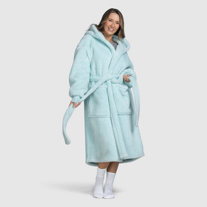 Woman wearing a light blue hooded bathrobe with pockets and a belt, smiling and holding the belt ends, standing barefoot with white socks.