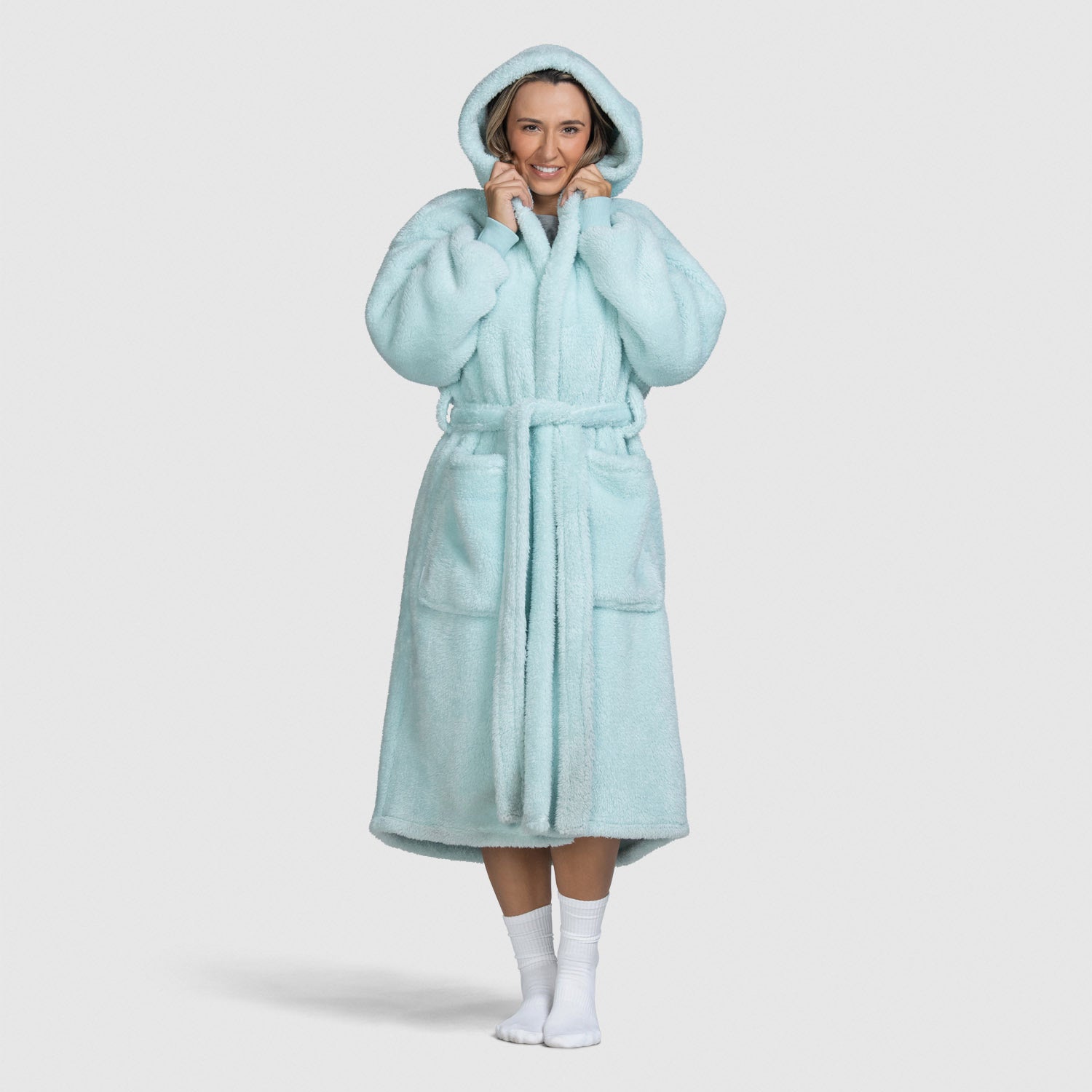 Woman wearing a light blue hooded bathrobe with pockets and belt, standing and holding the hood.