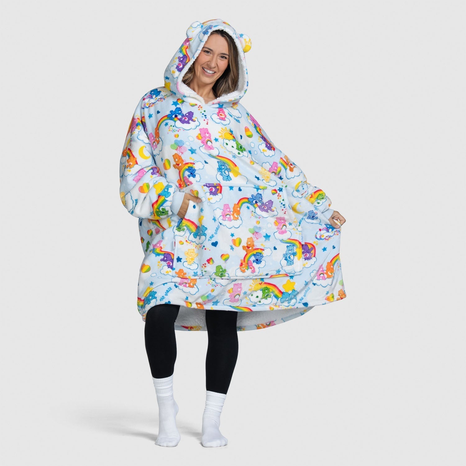 Woman wearing an oversized hoodie blanket with Care Bears characters, rainbows, and clouds pattern, smiling and posing with hands in pockets.