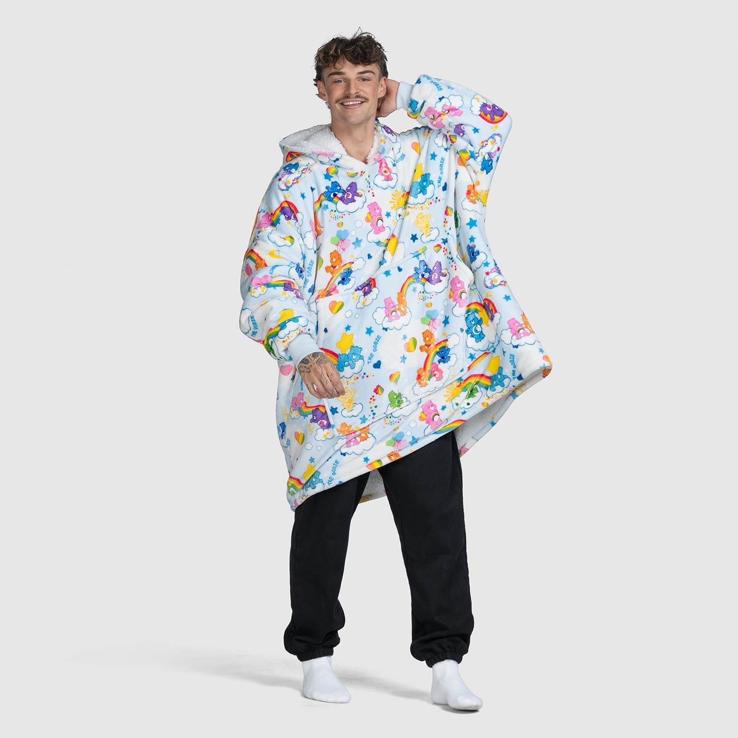 A smiling man wearing a colorful oversized hoodie with Care Bears and clouds pattern, standing against a plain background.