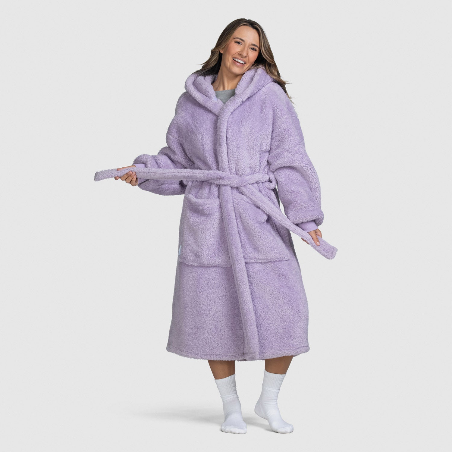 Woman wearing a lilac-colored plush hooded robe with a belt, smiling and adjusting the belt, standing barefoot with white socks.