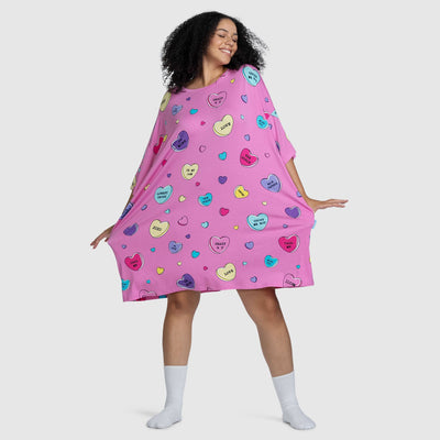 Woman wearing a loose pink dress with colorful heart shapes containing the words: CRAZY 4 U, LOVE, GHOST ME NOT, BE MINE, UR MY JAM, THE ODDIE, LOWKEY CRUSH, XOXO, CALL ME, MISS YOU, MIX SQUEEZE, BYE BYE.