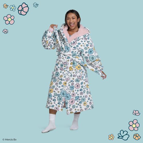 Woman smiling and posing in a white bathrobe with pink collar, decorated with Miffy characters and colorful flowers, against a blue background with floral illustrations.