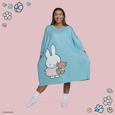 Woman smiling and posing in a blue dress featuring a large print of Miffy holding a teddy bear, with decorative flowers on a pink background.