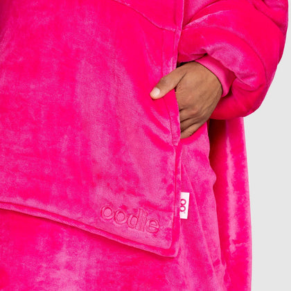 Hand in bright pink plush garment pocket with 'oodie' embroidered near hem and small white tag visible
