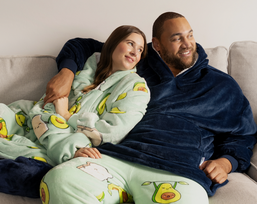 A couple sitting on a couch wearing cozy hooded blankets; the woman wears a light green avocado-themed blanket, and the man wears a navy blue blanket.