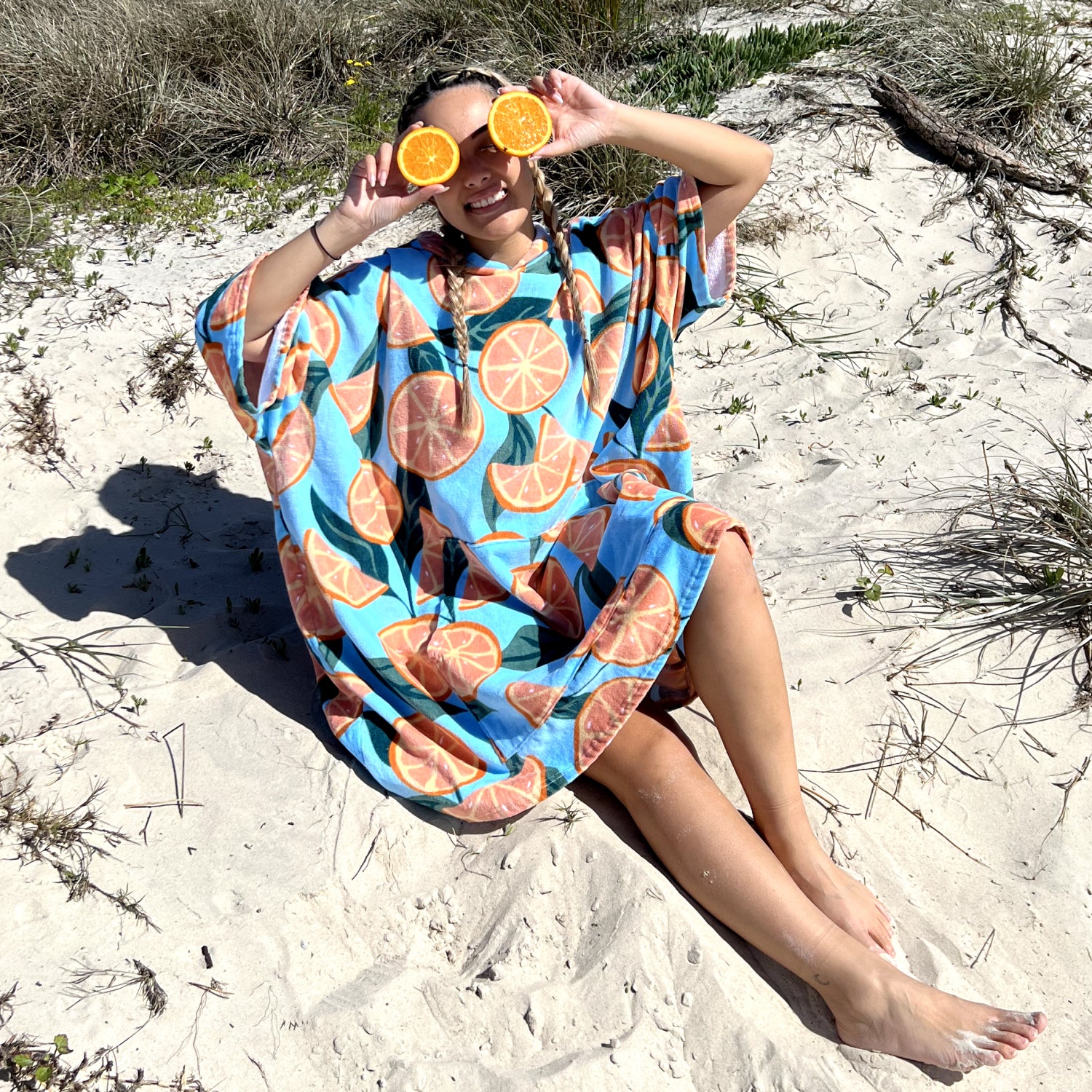 Person sitting on sand holding two orange halves over their eyes, wearing a blue garment with orange slice patterns.