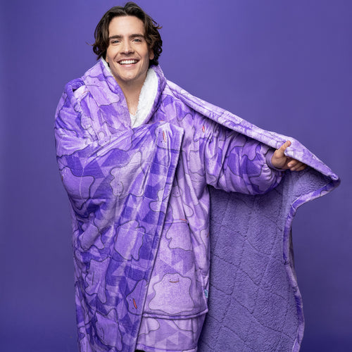 Man smiling and wrapped in a purple blanket patterned with multiple Ditto Pokémon faces, against a purple background.