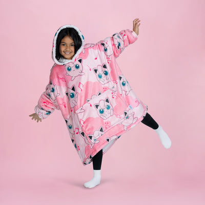 Child wearing a pink oversized hoodie blanket covered in Pokémon Jigglypuff faces, standing on one leg against a pink background.