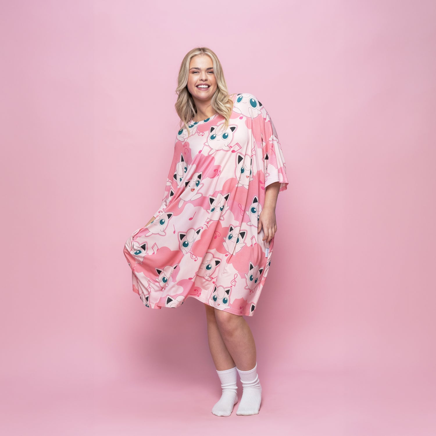 Woman wearing a pink dress covered with multiple Jigglypuff Pokémon prints, standing against a pink background.