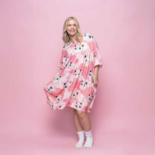 Woman wearing a pink dress covered with multiple Jigglypuff Pokémon prints, standing against a pink background.