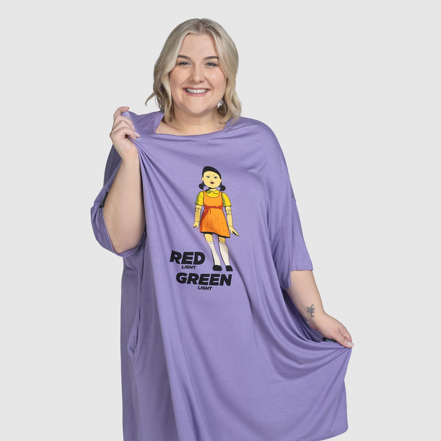 A smiling woman with blonde hair wearing a loose purple shirt featuring a graphic of a girl in an orange dress and the text 'RED LIGHT GREEN LIGHT'. Model Montana is 165cm tall and wears size S-XL. size: Montana is 165cm wearing a size S-XL
