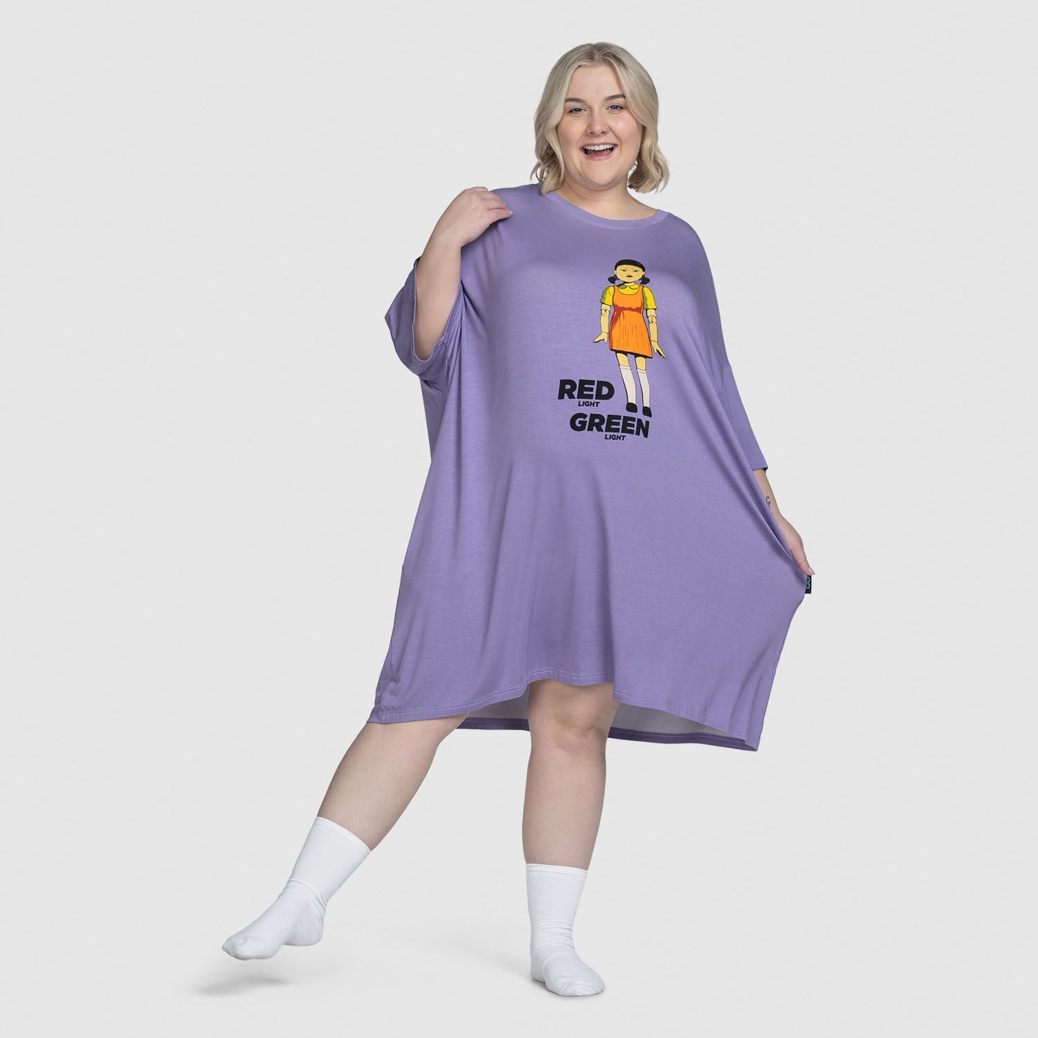 A smiling woman wearing a loose purple dress with a graphic of a woman in an orange dress and the words 'RED GREEN' on the front. Model is 5'5" wearing size S-XL. size: Montana is 5'5" wearing a size S-XL