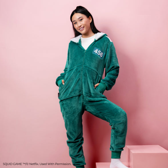 Young woman wearing a green Squid Game onesie with number 456 on chest, standing against a pink background with one foot on a pink step.