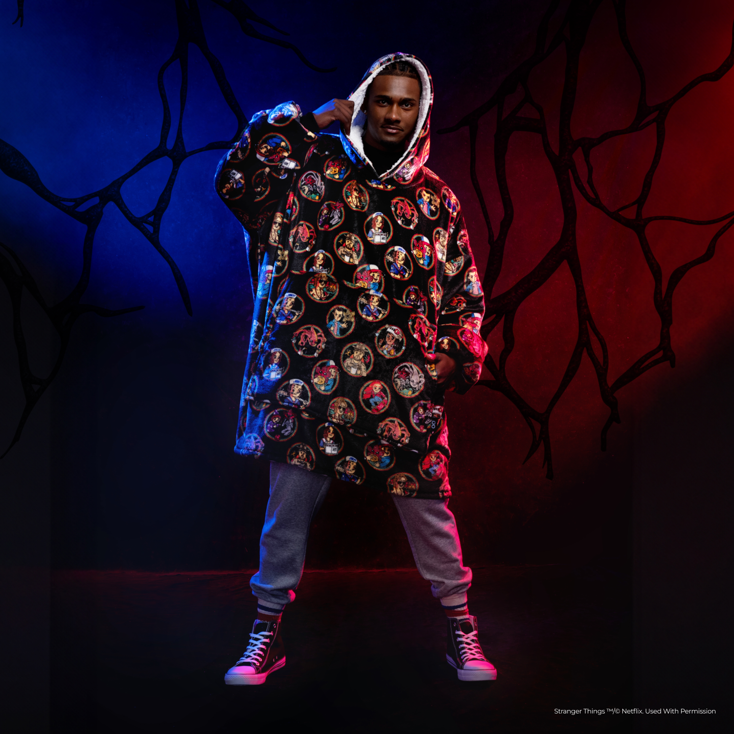 Man wearing a Stranger Things-themed oversized hoodie with character illustrations, standing against a dark background with red and blue lighting and branch-like patterns.