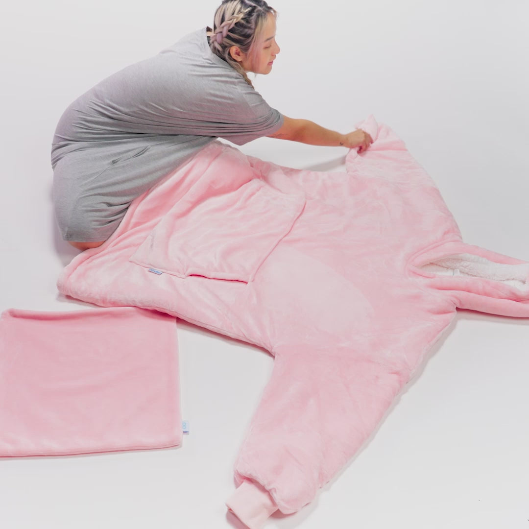 Person unfolding a large pink wearable blanket or onesie on a white background