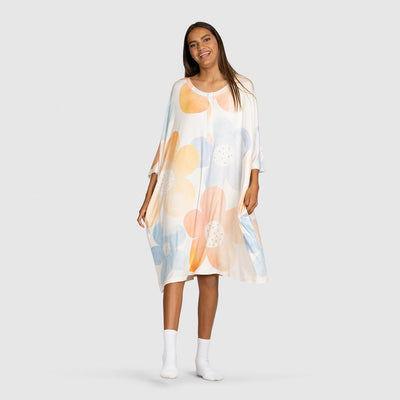 Woman wearing a loose, knee-length dress with large pastel orange, blue, and peach floral prints, standing and smiling against a plain background.