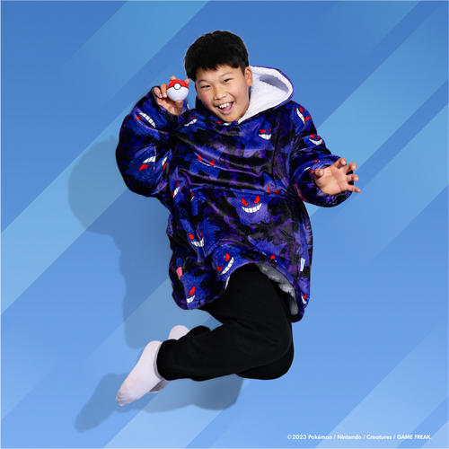 Boy jumping wearing a blue hoodie patterned with Pokémon Gengar faces, holding a Poké Ball in his right hand against a blue background