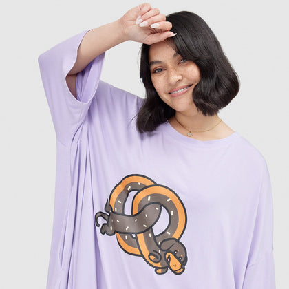 Smiling woman with short dark hair wearing a lavender shirt featuring a cartoon pretzel intertwined with a dachshund dog design
